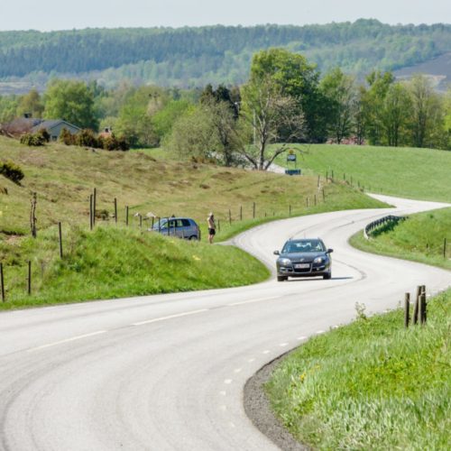 Brosarp: Documentary of Brosarps hills nature area. Car driving the winding road through the scenic landscape. Person standing roadside looking through a scope at car.